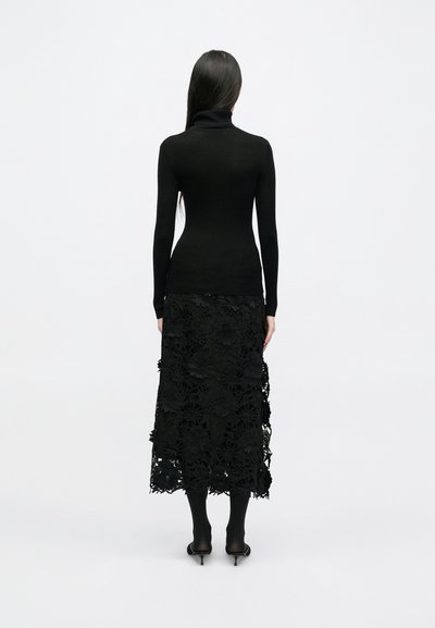 Un top negru cu guler înalt, smocks, asociat cu o fustă lungă din dantelă neagră cu modele florale și un tiv ondulat, purtat cu ciorapi subțiri negri și tocuri.