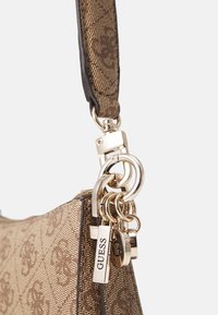 Ruskea kuvioitu käsilaukku, jossa on kultaiset yksityiskohdat, teksturoitu pinta ja useita riipuskoruja, mukaan lukien "GUESS"-tagi.