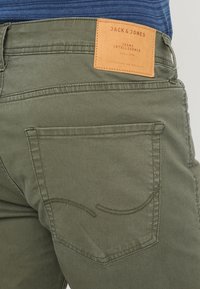 Olivgrüne Jack & Jones Jeans mit Gesäßtasche und braunem Leder-Label oberhalb des Bundes, getragen mit einem blauen Rippoberteil.