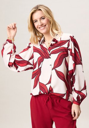 Lachende vrouw met blond haar, gekleed in een witte blouse met grote rode bladpatronen en rode broek met trekkoord, die één arm omhoog steekt.