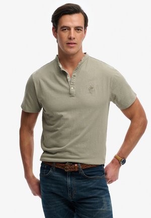 Mand iført lys olivengrøn kortærmet henley-shirt, flettet brunt læderbælte, blå jeans og armbåndsur, stående med den ene hånd på hoften.