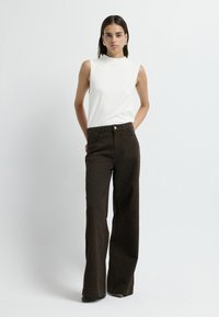 Top bianco senza maniche e jeans a zampa d'elefante marroni con decorazioni a pois, dotati di vita alta e tasche frontali. Tessuto dalla texture liscia.