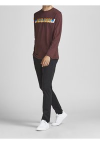 Man walking wearing a maroon long-sleeve Jack & Jones shirt, black pants, and white sneakers on a plain gray background.