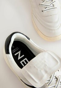 White sneakers with black accents, leather upper, textured design, contrasting laces, and Velcro strap. Interior labels visible.