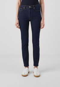 Tmavě modré džíny slim fit, s vysokým pasem, pěti kapsami a kontrastním oranžovým šitím. Nosené s bílými teniskami.