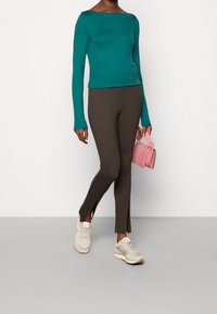 Grön långärmad topp med båthalsringning, kombinerad med bruna tajta byxor med sidospår. Bärs med beige sneakers och en rosa handväska.