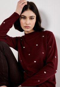 Pull bordeaux à col montant, orné de petits motifs en forme de cœur éparpillés dans des tons métalliques. Texture douce avec des poignets côtelés.
