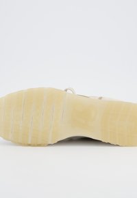 Sole tenisky s texturovanou gumovou základnou s vzorem. Barva je světlá tan s bílými akcenty. Obsahuje viditelné drážky a branding.