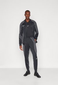 Juodas treniruočių kostiumas su baltais akcentais, turintis užtrauktuko striukę ir siaurėjantį kelnių siluetą. Pagamintas iš tamprios medžiagos, skirta komfortui ir judrumui.