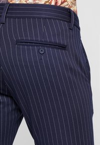 Pantalons rayés marine en tissu lisse. Dotés d'une poche arrière avec un bouton et d'une coupe ajustée. Les rayures verticales ajoutent de la texture.