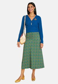 Blaues Wickel-Sweatshirt, grüne gemusterte Rock mit floralen Motiven, goldene Schuhe und eine metallische Schultertasche. Bemerkenswerte Details sind eine Brosche.