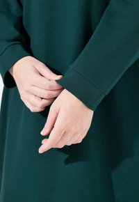 Prenda de manga larga en tela de color verde oscuro. Presenta un corte holgado con mangas ajustadas. Textura suave, diseño minimalista y sin patrones visibles.