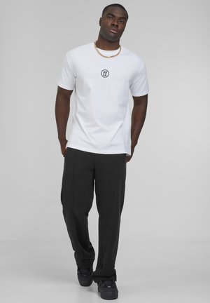 Camiseta blanca de cuello redondo con un pequeño logo negro en el pecho, combinada con pantalones de chándal negros que cuentan con costuras visibles y bolsillos.