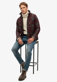 Rød og sort ternet flannel skjorte over en creme sweater, parret med blå jeans og brune snørebåndsstøvler, siddende på en sort skammel.