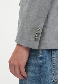 Χέρι χαλαρό στο πλάι, φορώντας ανοιχτό γκρι σακάκι με τέσσερις κουμπιά στους καρπούς και μπλε τζιν παντελόνι.