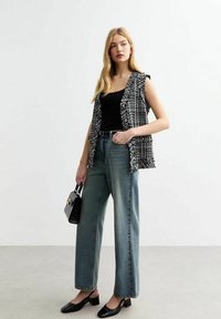 Schwarz-weißer Tweed-Weste über einem schwarzen Tanktop, kombiniert mit hellen, weit geschnittenen Jeans und schwarzen Blockabsatzschuhen. Kleine Handtasche inklusive.