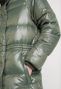 Mano en el bolsillo de una brillante chaqueta acolchada de invierno verde oliva con detalles en la cintura y los puños elásticos.
