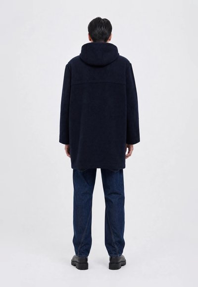 Mann trägt einen marineblauen Kapuzenmantel, dunkelblaue Jeans und schwarze Schuhe, steht mit dem Rücken zu einem schlichten hellen Hintergrund.