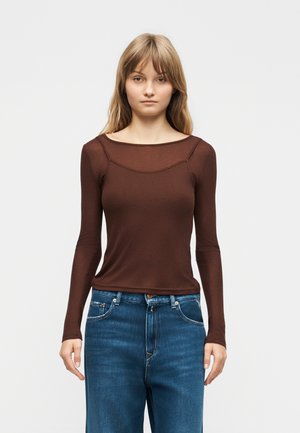 Mladá žena s rovnými blond vlasmi, oblečená v priliehavom hnedom tričku s dlhým rukávom a modrých džínsach, stojaca pred jednoduchým svetlým pozadím.