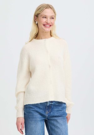 Femme souriante aux longs cheveux blonds portant un pull blanc boutonné et un jean bleu devant un fond clair uni.