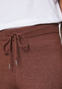 Hnědé pletené joggery s žebrovaným pasem, vybavené šňůrkou na stažení a stříbrnými kovovými špičkami, detailní záběr na horní část.