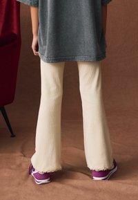 Beige weit ausgestellte Hosen mit gerippter Textur, kombiniert mit lila Sneakers, die kontrastierende weiße Akzente haben, getragen unter einem lockeren grauen Oberteil.