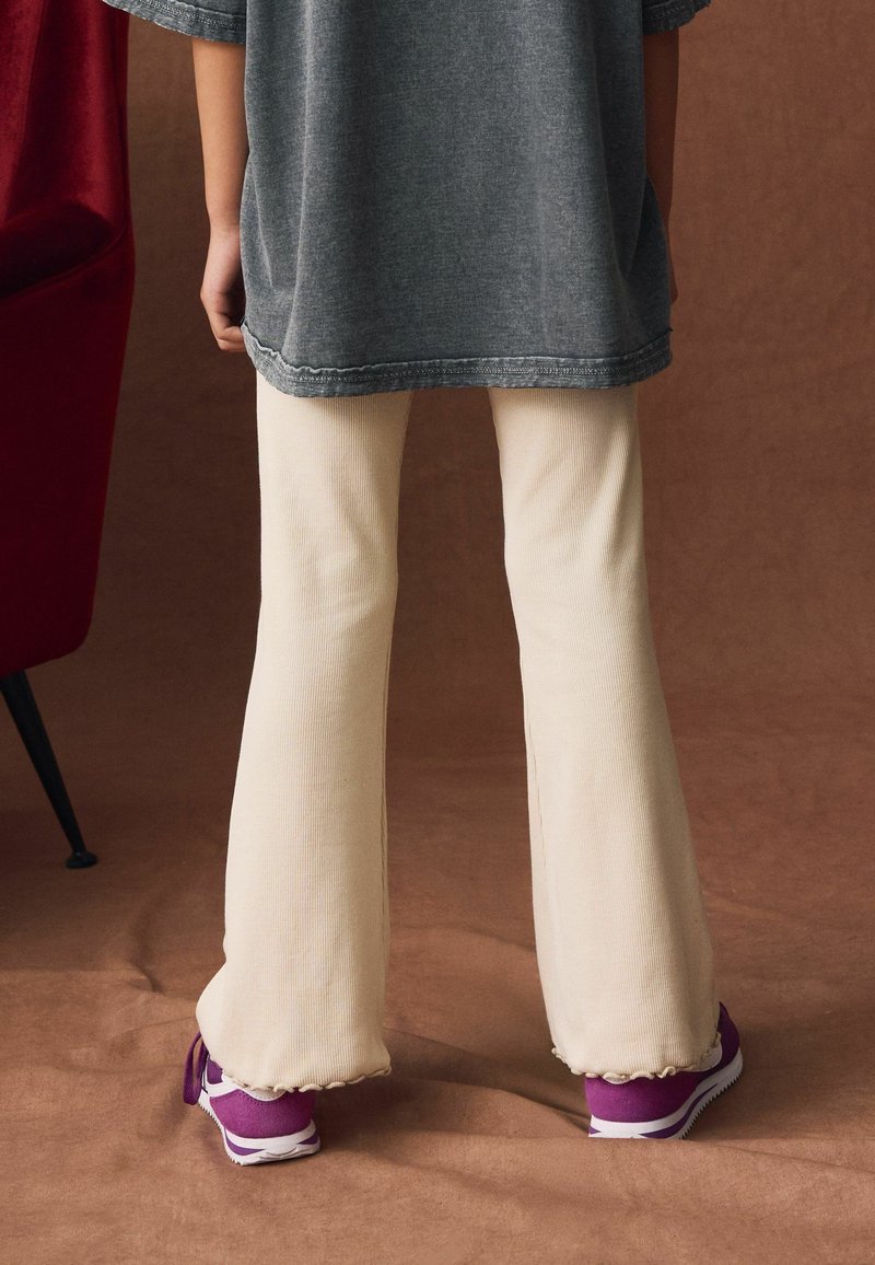 Beige weit ausgestellte Hosen mit gerippter Textur, kombiniert mit lila Sneakers, die kontrastierende weiße Akzente haben, getragen unter einem lockeren grauen Oberteil.
