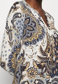 Blusa con motivo caratterizzata da disegni floreali blu, oro e bianchi. Tessuto leggero con scollatura a V e lunghe maniche a sbuffo.