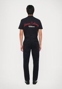 Czarna koszulka z czerwoną haftowaną grafiką "Armani Exchange Milano" na plecach, w zestawie z ciemnymi spodniami i czarnymi butami.