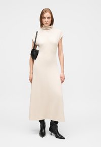 Μπεζ εφαρμοστό μίντι φόρεμα με ψηλό λαιμόκοψη cowl και κοντά μανίκια, σε συνδυασμό με μαύρες μπότες μέχρι το γόνατο και μια μικρή μαύρη τσάντα ώμου.