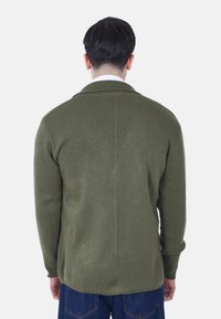 Cardigan lavorato a maglia verde oliva con texture a coste, colletto a scialle, maniche lunghe e cucitura posteriore diritta. Indossato con jeans di denim blu.