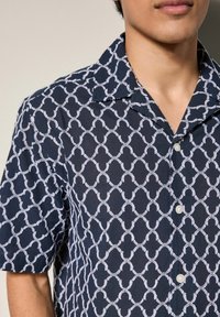 Cămașă cu mânecă scurtă, de culoare bleumarin, având un model geometric alb, cu închidere prin butoni și guler clasic, realizată dintr-un material neted.