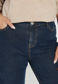 Jeans de mezclilla azul oscuro con costuras naranjas en contraste, cierre de botón metálico y bolsillos frontales. Textura suave, corte casual.