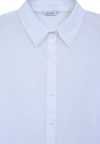 Chemise blanche boutonnée en tissu texturé. Elle présente un col classique et cinq boutons sur le devant, avec une petite étiquette à l'intérieur.