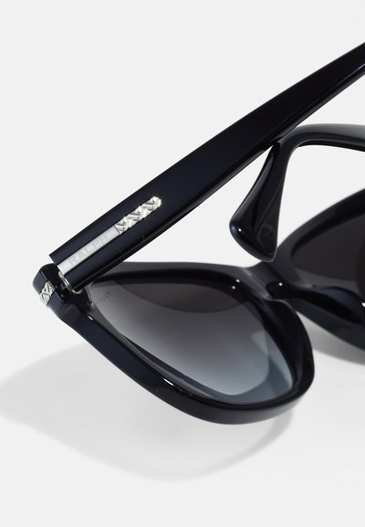 Ochelari de soare negri cu lentile în degrade întunecat, cadru unghiular și accente argintii strălucitoare pe brațe, prezentând o textură plastică elegantă.
