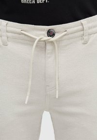 Beige corduroy pants with a ribbed texture, featuring a drawstring closure and a branded black button at the waist.