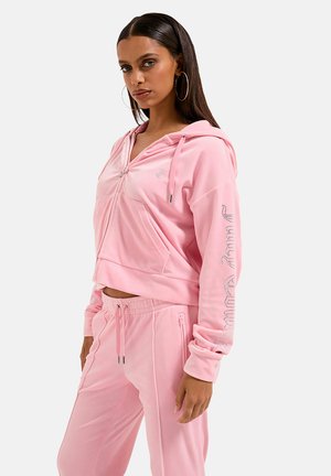 Vrouw met lang donker haar, grote ronde oorbellen en een roze zip-up hoodie met bijpassende roze joggingbroek, staand in zijaanzicht.