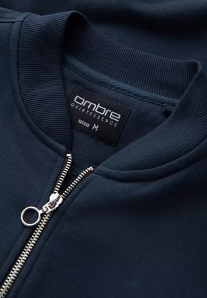 Granatowy sweatshirt z ściągaczami na mankietach i kołnierzu, z metalowym zamkiem błyskawicznym oraz czarną metką z napisem "ombre quintessence" w rozmiarze M.