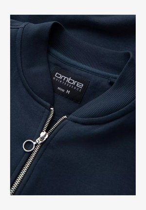 Navyblauwe sweatshirt met ribbelmanchetten en kraag, voorzien van een zilveren rits en een zwart label met "ombre quintessence" en maat M.