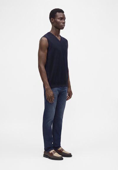 Homme debout portant un pull sans manches bleu marine foncé, un jean bleu et des chaussures habillées bicolores marron et beige sur fond blanc.