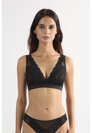 Kvinde iført sort blonder bralette og matchende blonder undertøj, stående mod en ensfarvet lys baggrund, vendt fremad.