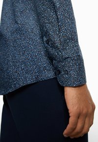 Marineblaues Hemd mit kleinem, mehrfarbigem Blumenmuster, Knopfmanschetten und einer glatten Textur, kombiniert mit dunkler Hose.