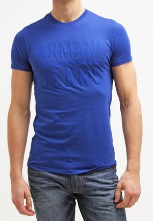 Mężczyzna ubrany w dopasowaną niebieską koszulkę Armani Jeans i wyblakłe niebieskie dżinsy, stojący na tle jednolitego białego tła.