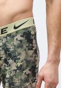 Κοντινό πλάνο ενός ατόμου που φοράει σορτς Nike με πράσινο και μαύρο καμουφλάζ, με μπεζ ζώνη και ορατά λογότυπα Nike.