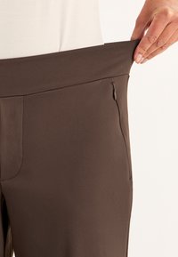Bruine broek met een gladde textuur, elastische tailleband en een zijzak met ritssluiting. De stof lijkt lichtgewicht en rekbaar te zijn.