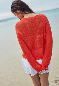 Pull en tricot rouge avec un motif en zigzag, doté d'un décolleté festonné et d'un tissage ouvert, porté par-dessus une robe blanche à ourlet brodé.