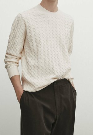 Person wearing a cream cable-knit sweater with long sleeves tucked into black high-waisted trousers, hands in pockets, against a plain light background.