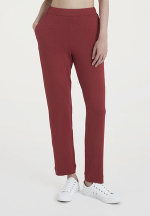 Persona con pantaloni rossi a gamba dritta con vita elastica e scarpe da ginnastica bianche, una mano in tasca e l'altra appoggiata di lato.