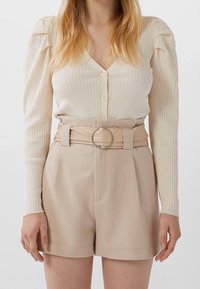 Cremefarbener gerippter Cardigan mit Knopfverschluss, kombiniert mit hochtaillierten beigen Shorts, die einen Gürtel mit einer silbernen runden Schnalle haben.