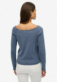 Top azul de manga larga y off-the-shoulder con una textura suave, presenta un dobladillo redondeado y mangas raglán, llevado con pantalones blancos.